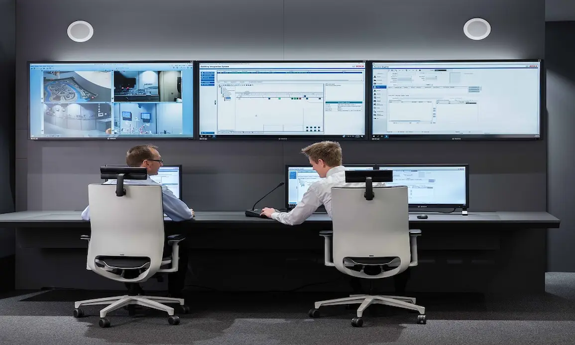 Two operators in a security control room monitoring multiple integrated security systems including CCTV, access control, intrusion detection and alarm systems