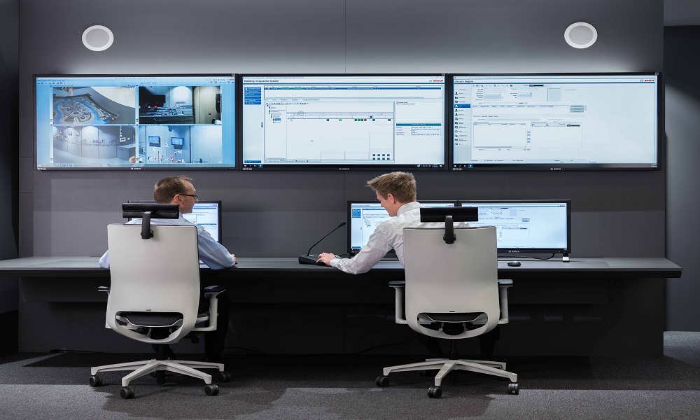 Two operators in a security control room monitoring multiple integrated security systems including CCTV, access control, intrusion detection and alarm systems