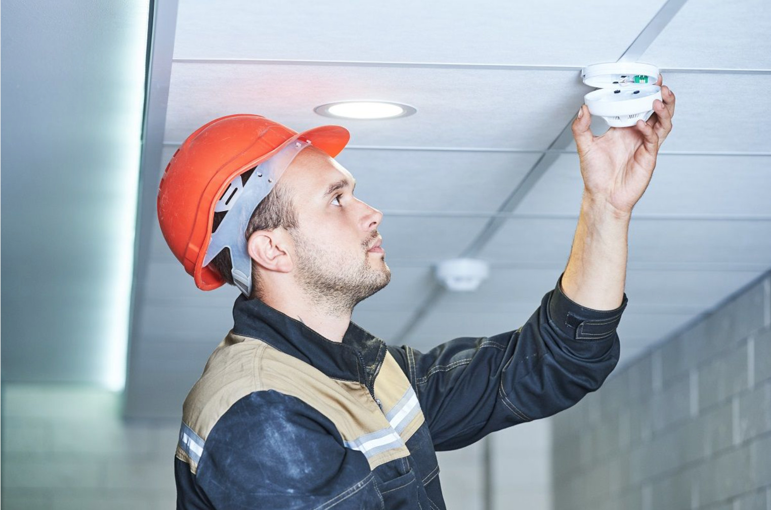 Technician installing white smoke detector on ceiling for commercial fire safety system setup.